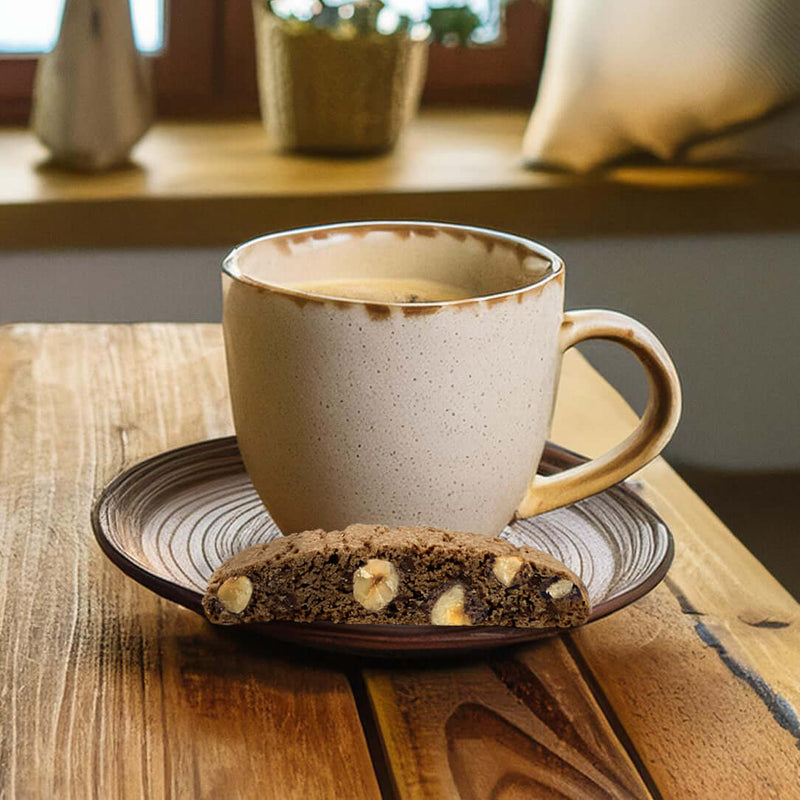 A beige ceramic mug of coffee sits on a matching saucer holding a True Delicious Chocolate Hazelnut Biscotti, made with Oregon hazelnuts. The set rests on a wooden table with soft light and decor in the background.