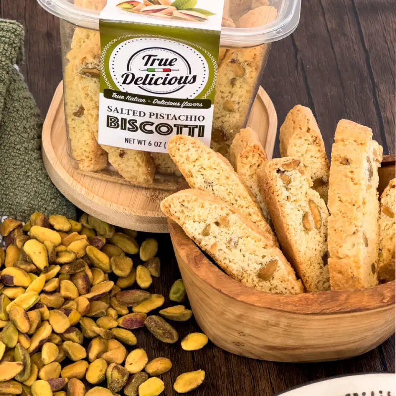 Bowl of salted pistachio biscotti with packaging labeled 'True Delicious' on a wooden surface.