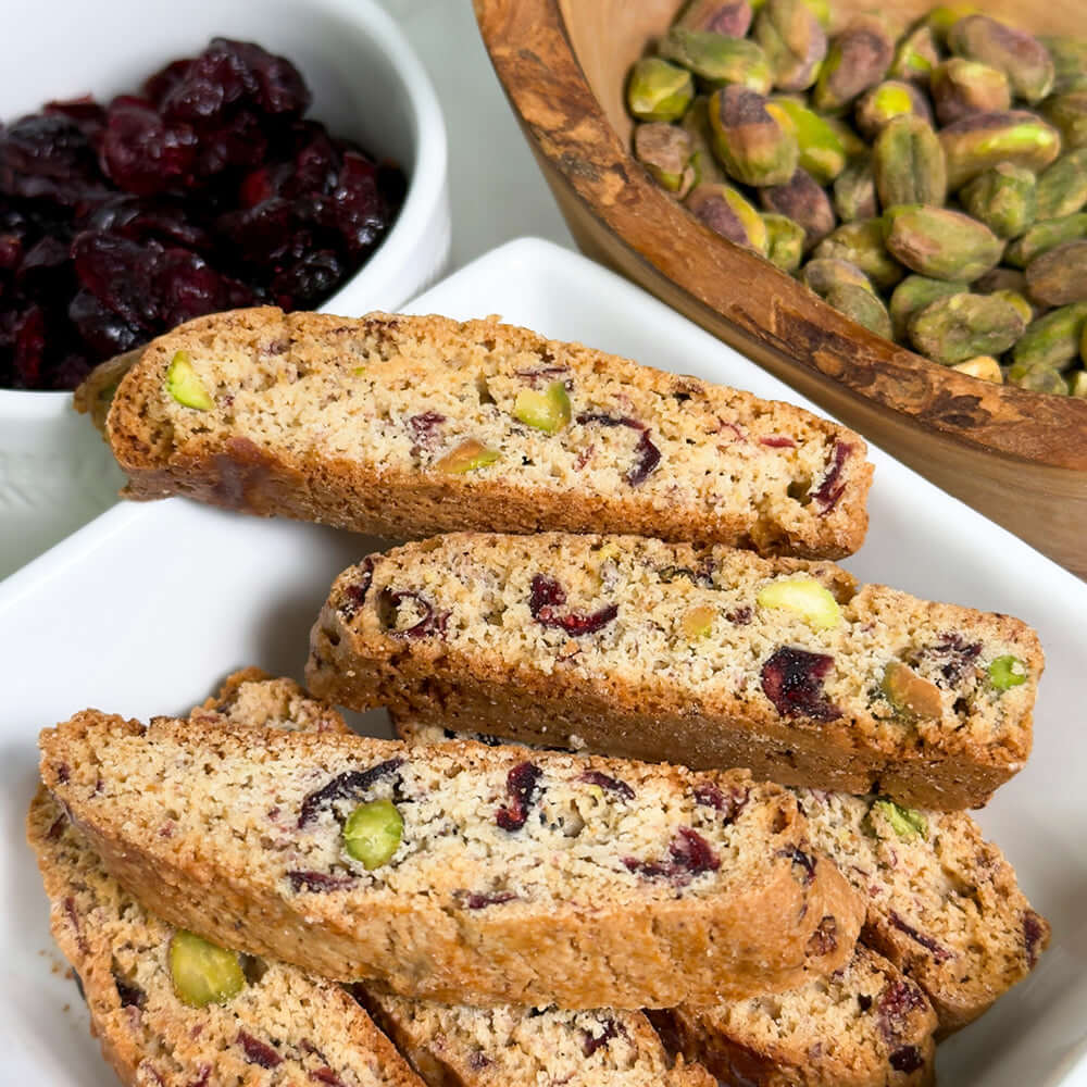 A plate of True Delicious Cranberry Pistachio Biscotti - Limited Edition, featuring visible pistachios and dried cranberries, sits beside bowls of dried cranberries and shelled pistachios—a treat for handcrafted biscotti fans.