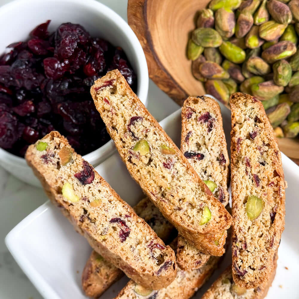 A plate of True Delicious Cranberry Pistachio Biscotti - Limited Edition with green pistachios and red cranberries, beside a bowl of dried cranberries and a wooden bowl of shelled pistachios.