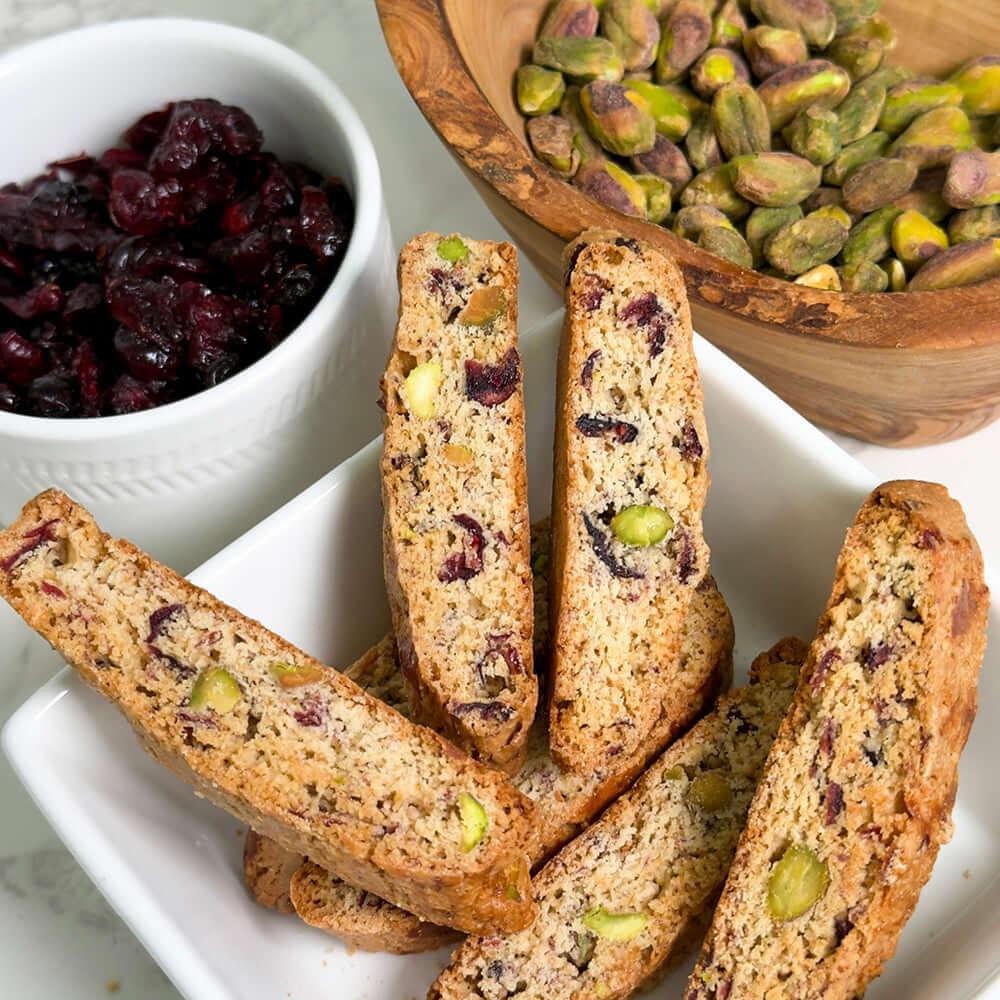 A bowl of True Delicious Cranberry Pistachio Biscotti - Limited Edition sits beside dried cranberries and pistachios, beautifully arranged on a marble surface to showcase this handcrafted treat.