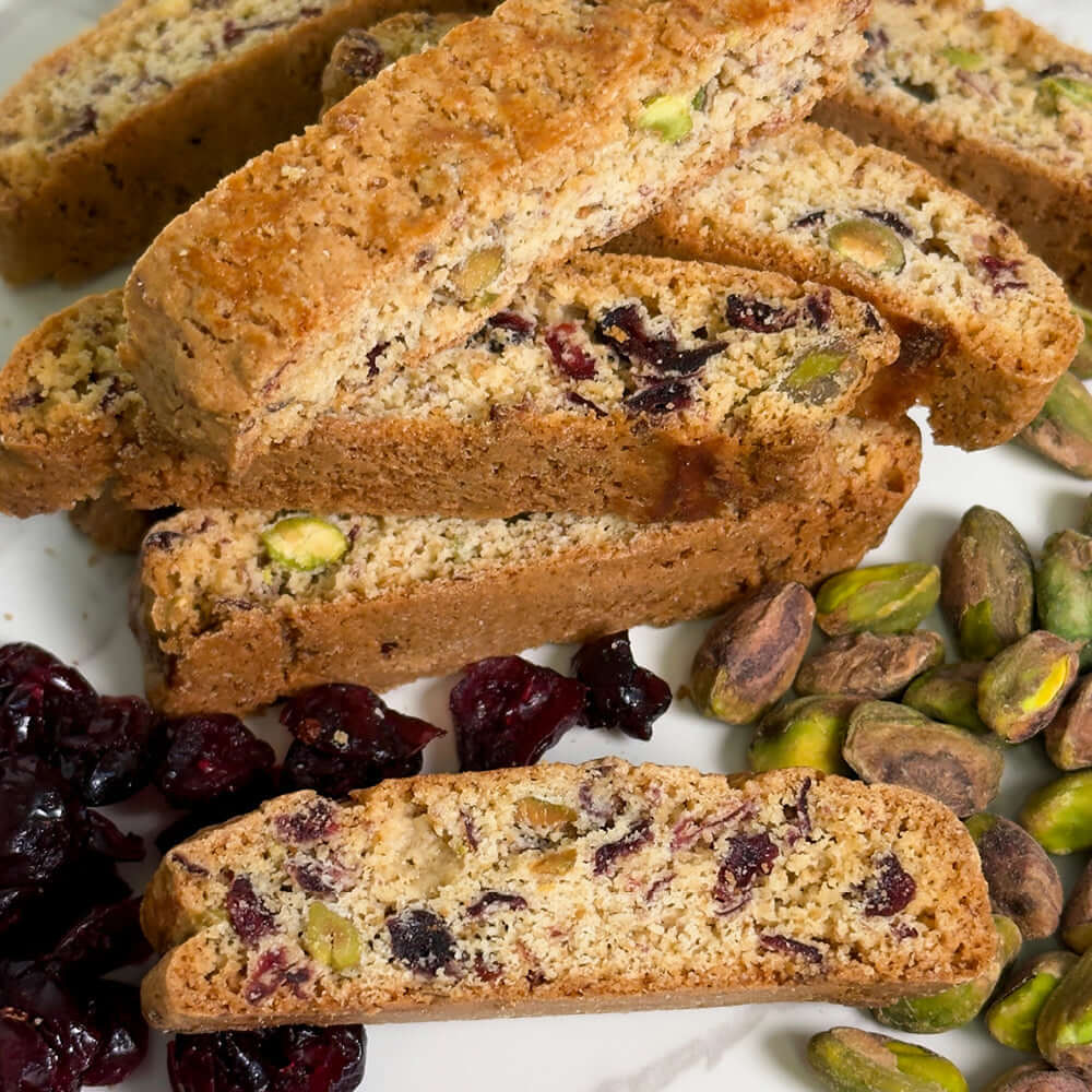 A stack of True Delicious Cranberry Pistachio Biscotti - Limited Edition with visible pistachios and cranberries, surrounded by scattered nuts and fruit on a white surface.