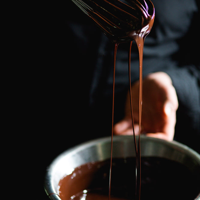 A close-up of a hand holding a whisk with True Delicious Chocolate Covered Almond Biscotti Bites being mixed in a metal bowl, set against a dark background.