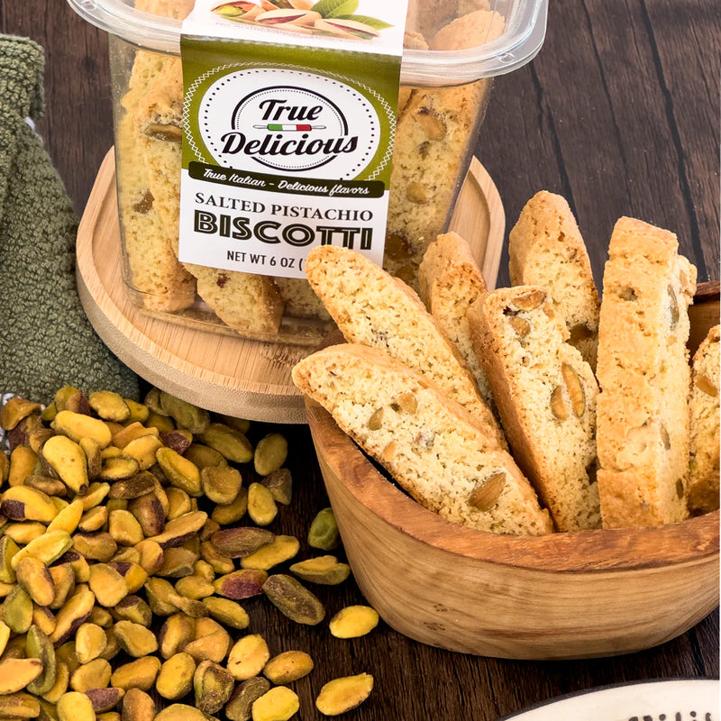 A container and wooden bowl filled with True Delicious Bundle of the Month — The Sweet Deal non-GMO Italian biscotti are displayed next to a pile of shelled pistachios on a wooden surface, with a green towel in the background.