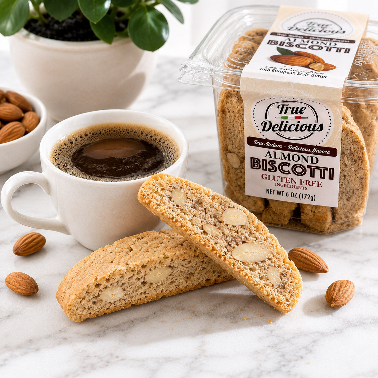 A cup of black coffee sits next to handcrafted biscotti, loose almonds, and a clear container labeled True Delicious Gluten Free Ingredients Almond Biscotti. A potted plant is in the background, highlighting this inviting gluten-free snack.