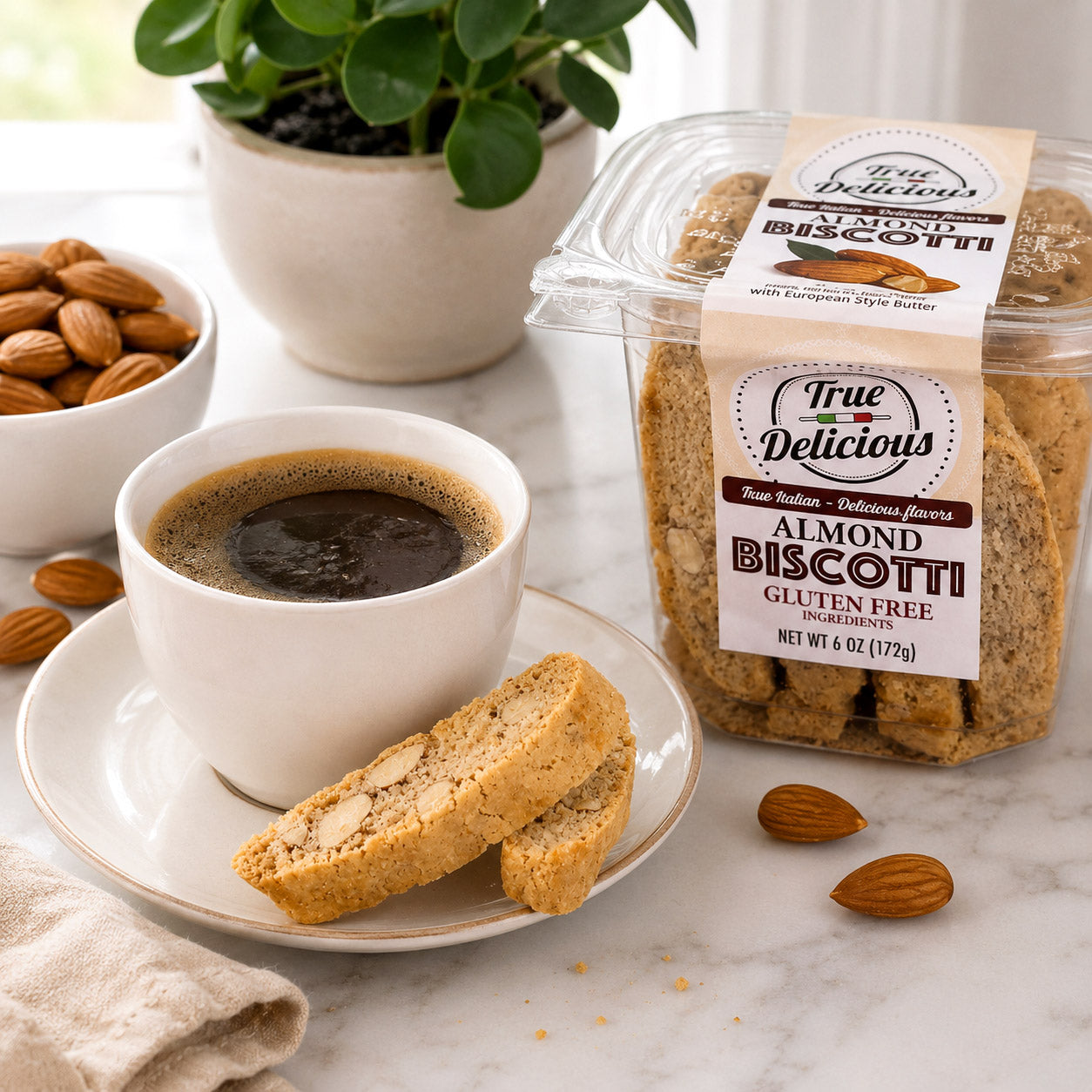 A cup of black coffee with two True Delicious Gluten Free Ingredients Almond Biscotti on a plate, a container of True Delicious biscotti, almonds, and a potted plant in the background on a light-colored table.