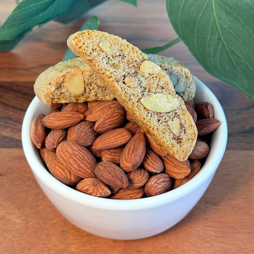 A white bowl filled with whole almonds and two pieces of True Delicious Gluten Free Ingredients Almond Biscotti sits on a wooden surface with green leaves in the background.