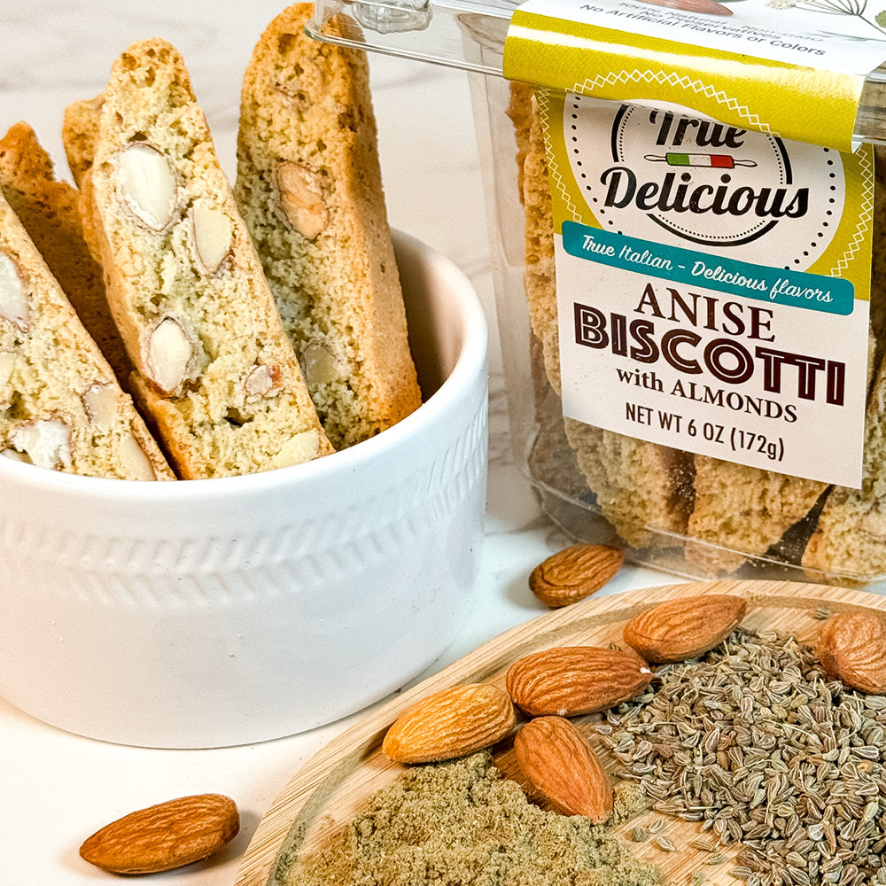 A white bowl filled with True Delicious Anise Biscotti with Almonds sits next to a clear package of the product, while whole almonds and anise seeds are scattered on a wooden surface, highlighting authentic Italian baking.