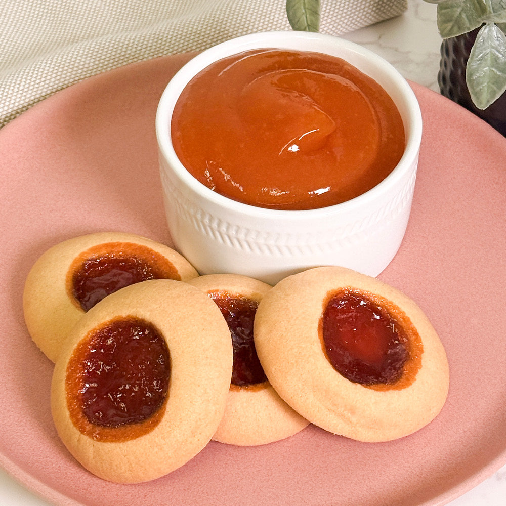 A pink plate holds four True Delicious Apricot Jam Cookies—shortbread rounds filled with red jam—next to a white bowl of smooth orange apricot jam, with green leaves and a beige cloth in the background.