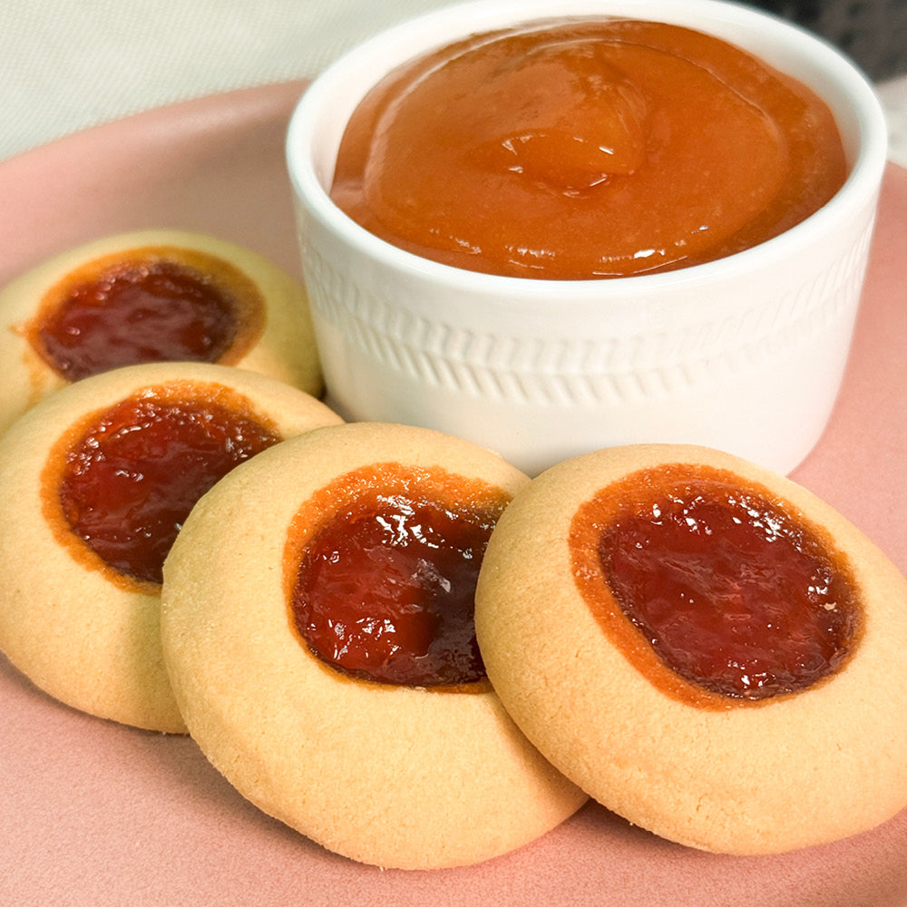 Four True Delicious Apricot Jam Cookies with buttery shortbread and apricot jam centers are arranged on a pink plate beside a white ramekin of orange spread.