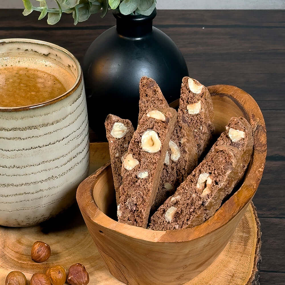 A rustic wooden bowl filled with True Delicious Chocolate Hazelnut Biscotti, made with Oregon hazelnuts and Guittard chocolate chips, sits on a wooden surface by a coffee mug, with a black vase and leafy plant in the background.