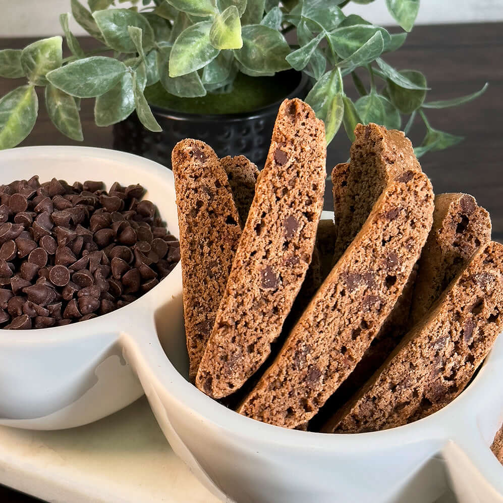 A white bowl holds several True Delicious Double Chocolate Biscotti, standing upright beside a bowl of chocolate chips, with a green leafy plant in the background.