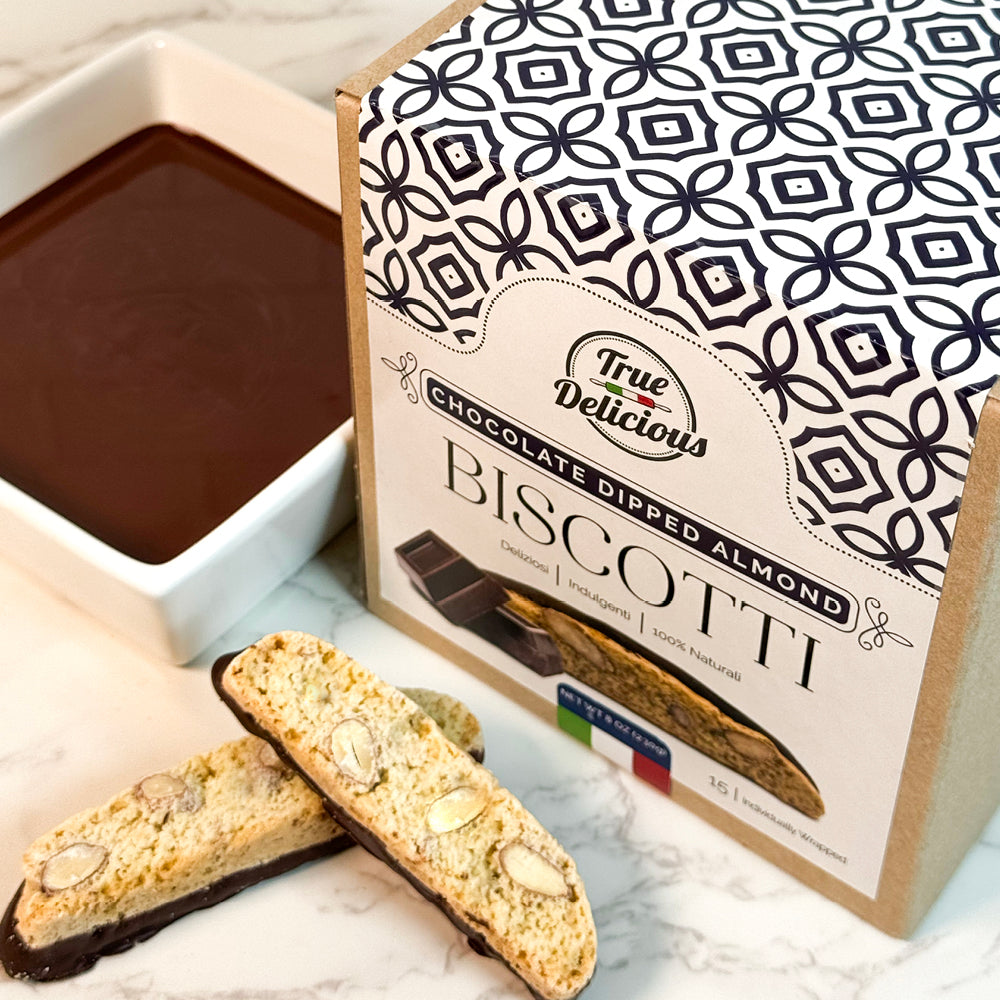 A box of True Delicious Elegant Box Chocolate Dipped Almond Biscotti sits beside two almond biscotti pieces and a white bowl of melted chocolate on a marble surface. Each biscotti is individually wrapped for freshness.