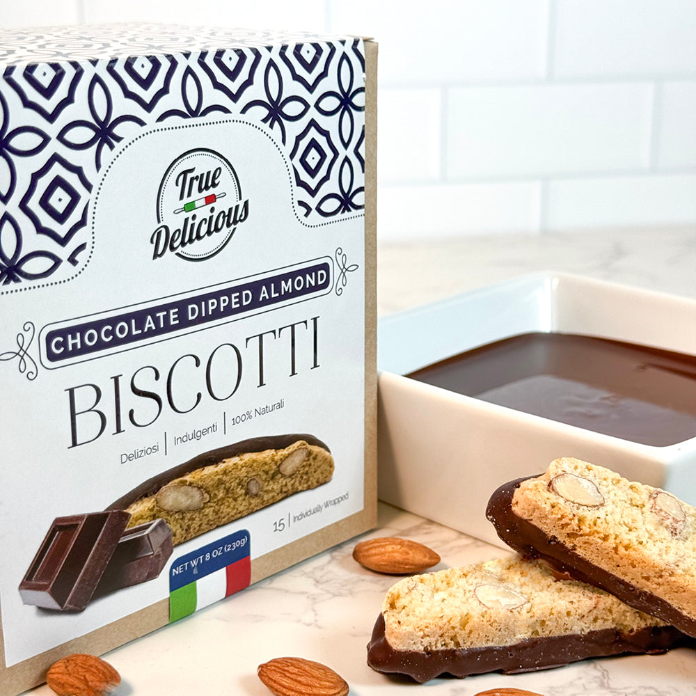 An Elegant Box of True Delicious Chocolate Dipped Almond Biscotti, individually wrapped, sits beside a dish of melted chocolate, with two chocolate-dipped biscotti and whole almonds arranged in front.