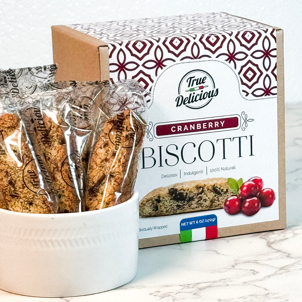 A box of True Delicious Elegant Box Cranberry Biscotti Individually Wrapped sits behind a white bowl filled with the treats on a marble countertop. The Italian packaging features elegant patterns and cranberry artwork.