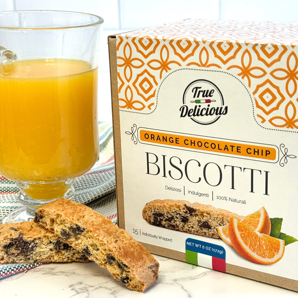 A glass of orange juice, a biscotti, and the True Delicious Elegant Box Orange Chocolate Chip Biscotti Individually Wrapped on a white surface. The box shows Italian biscotti and orange slices, with a patterned cloth in the background.
