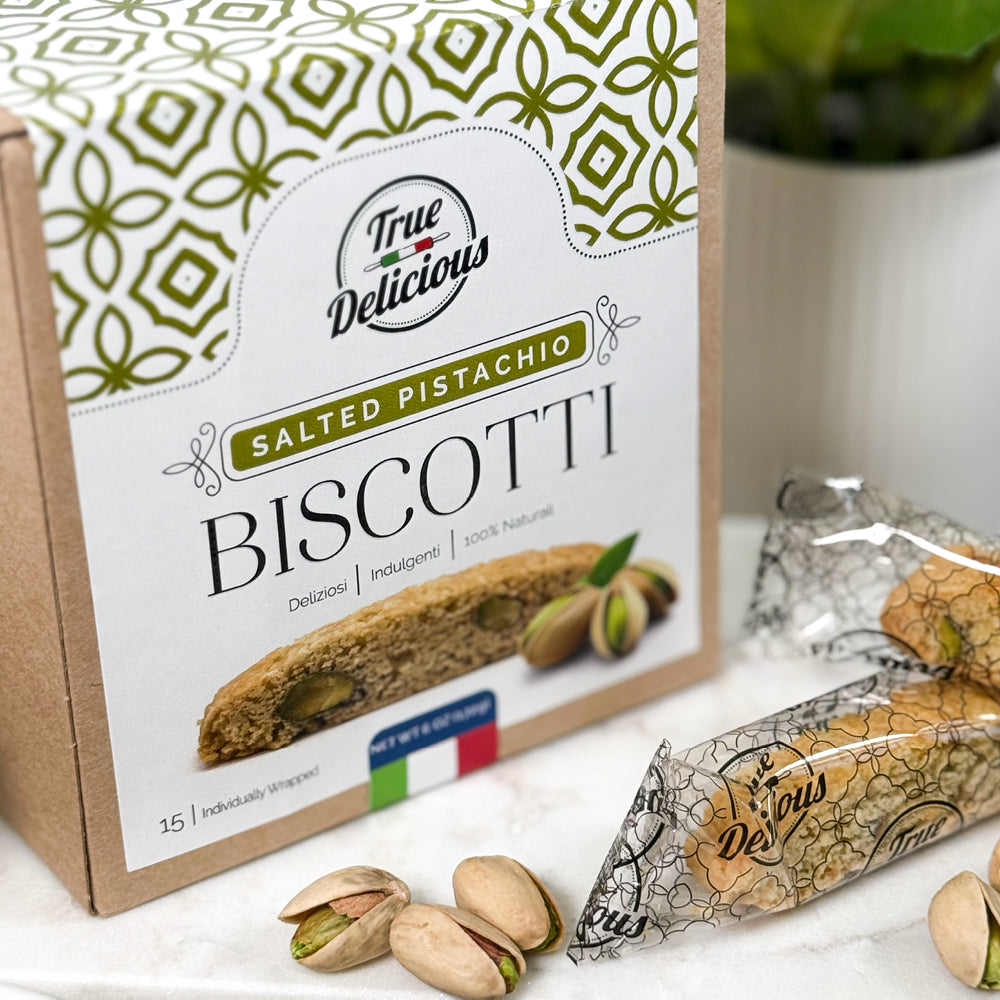 A box of True Delicious Elegant Box Salted Pistachio Biscotti Individually Wrapped shows biscotti and pistachios on the packaging, with individually wrapped biscotti and pistachios displayed before a blurred green plant background.