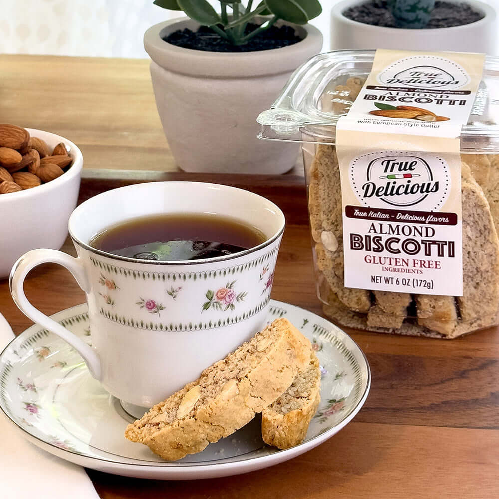 A cup of tea on a saucer sits beside two handcrafted True Delicious Gluten Free Ingredients Almond Biscotti, with a labeled container and a bowl of almonds on a wooden table. Two potted plants are in the background.