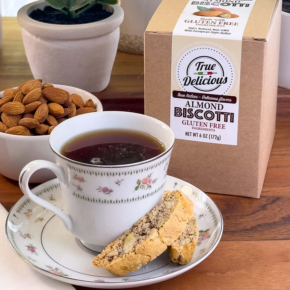 A cup of black coffee on a floral saucer with an almond biscotti, a bowl of California Almonds, and a box of True Delicious Gluten Free Ingredients Almond Biscotti - Individually Wrapped sit on a wooden table with potted plants in the background.