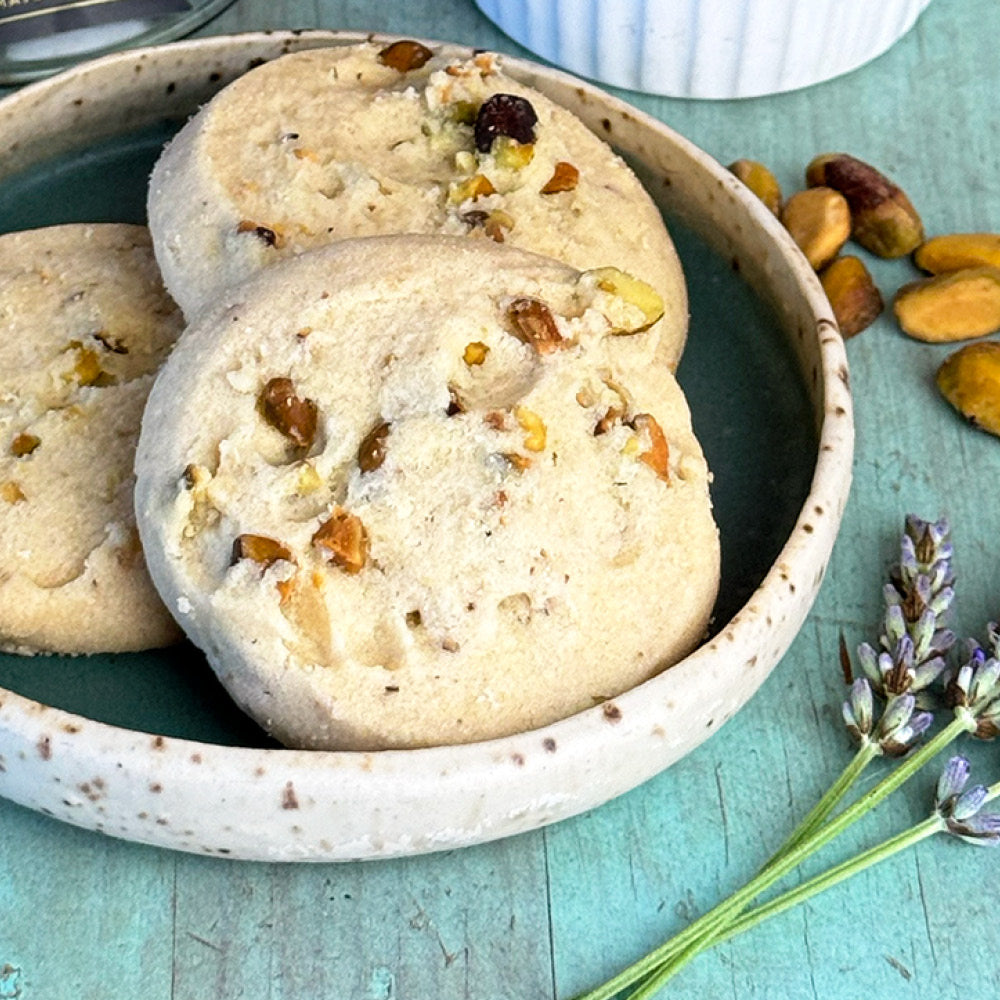 A speckled ceramic bowl holds three Pistachio Shortbread Cookies by True Delicious Biscotti, each topped with chopped pistachios. Lavender sprigs and scattered pistachios lie on a turquoise surface beside the bowl.