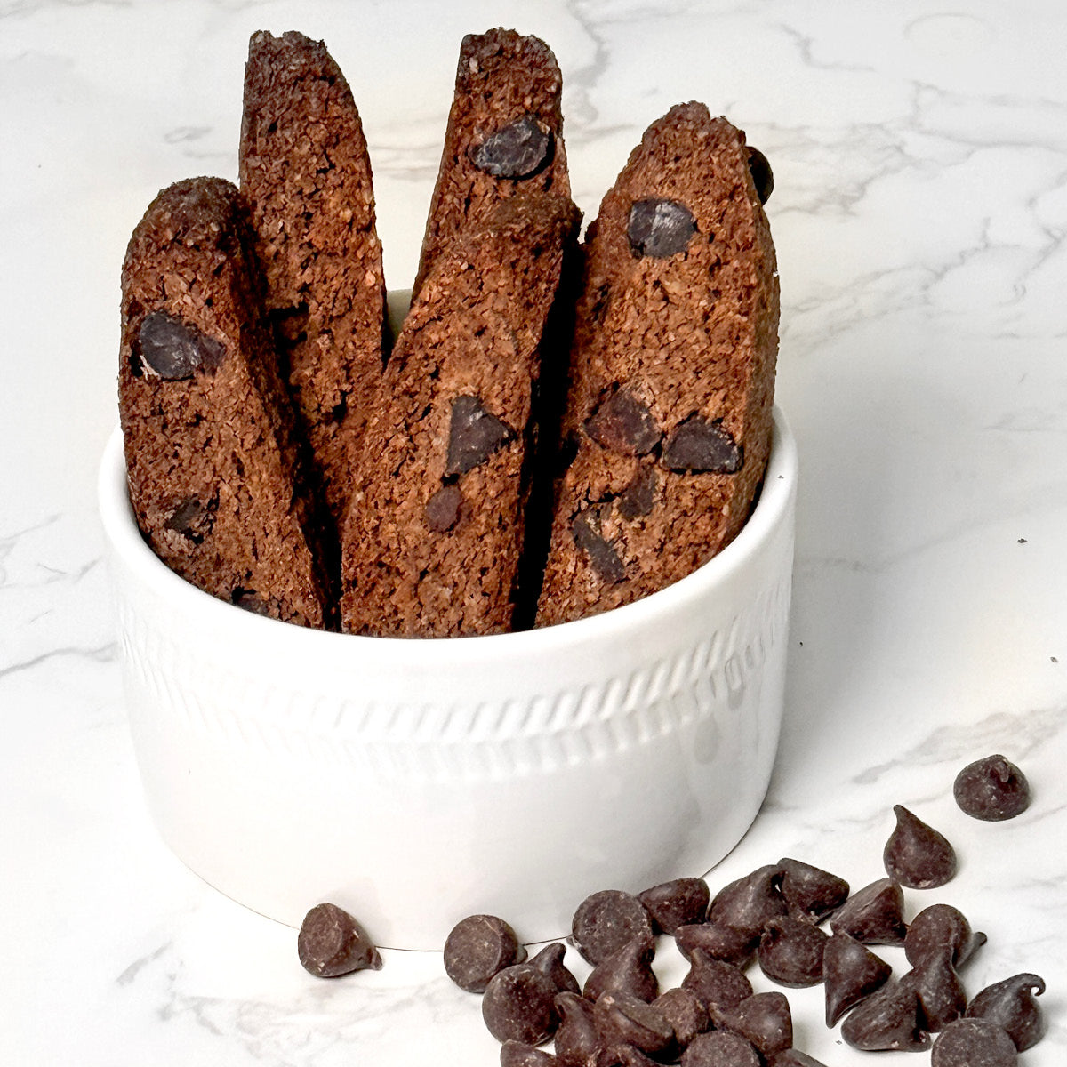 Four True Delicious Sugar Free Chocolate Biscotti stand upright in a white textured ceramic bowl, with chocolate chips scattered on the white marble surface beside the bowl.